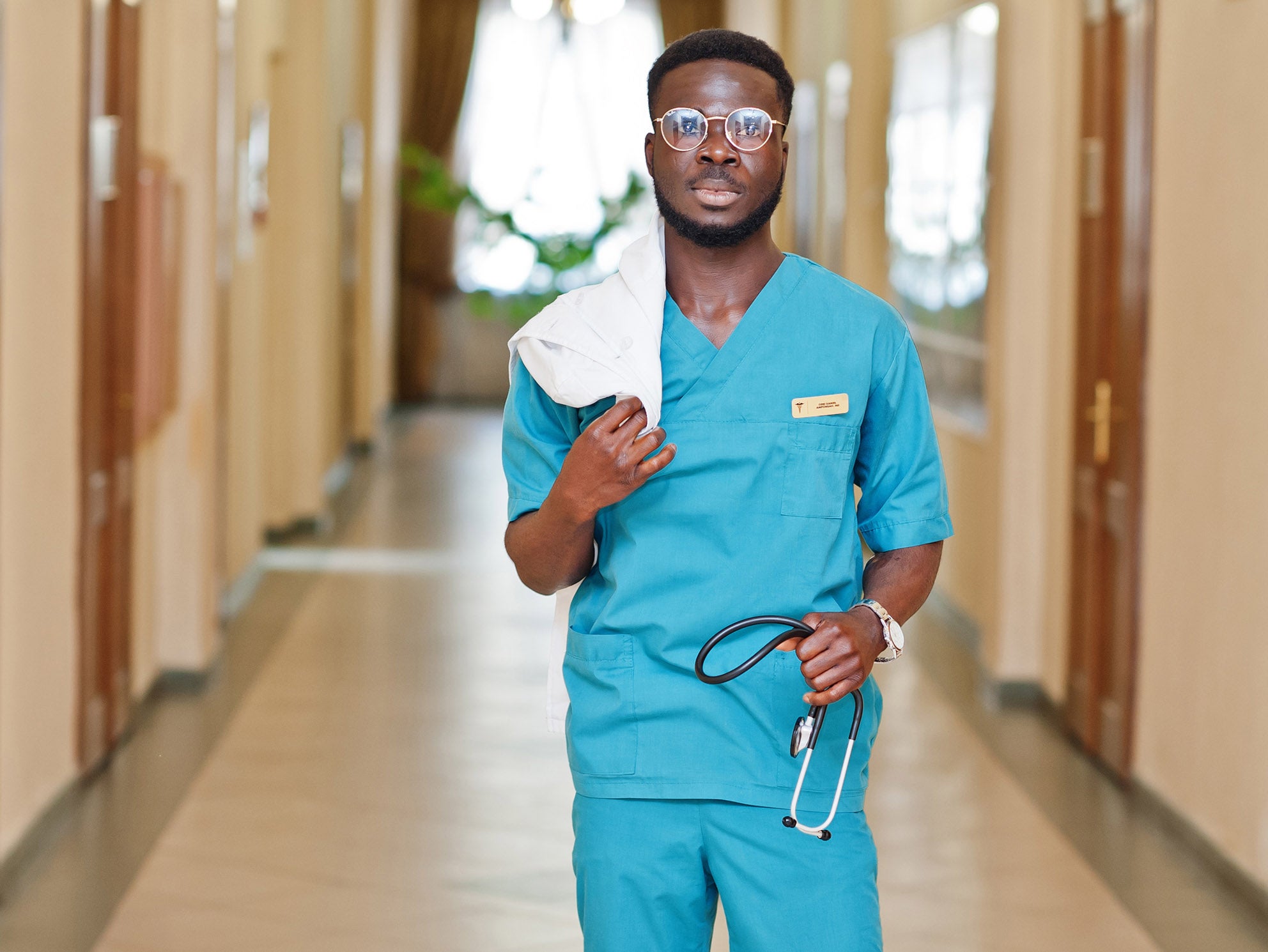 A man in scrubs holding a white coat and stethoscope